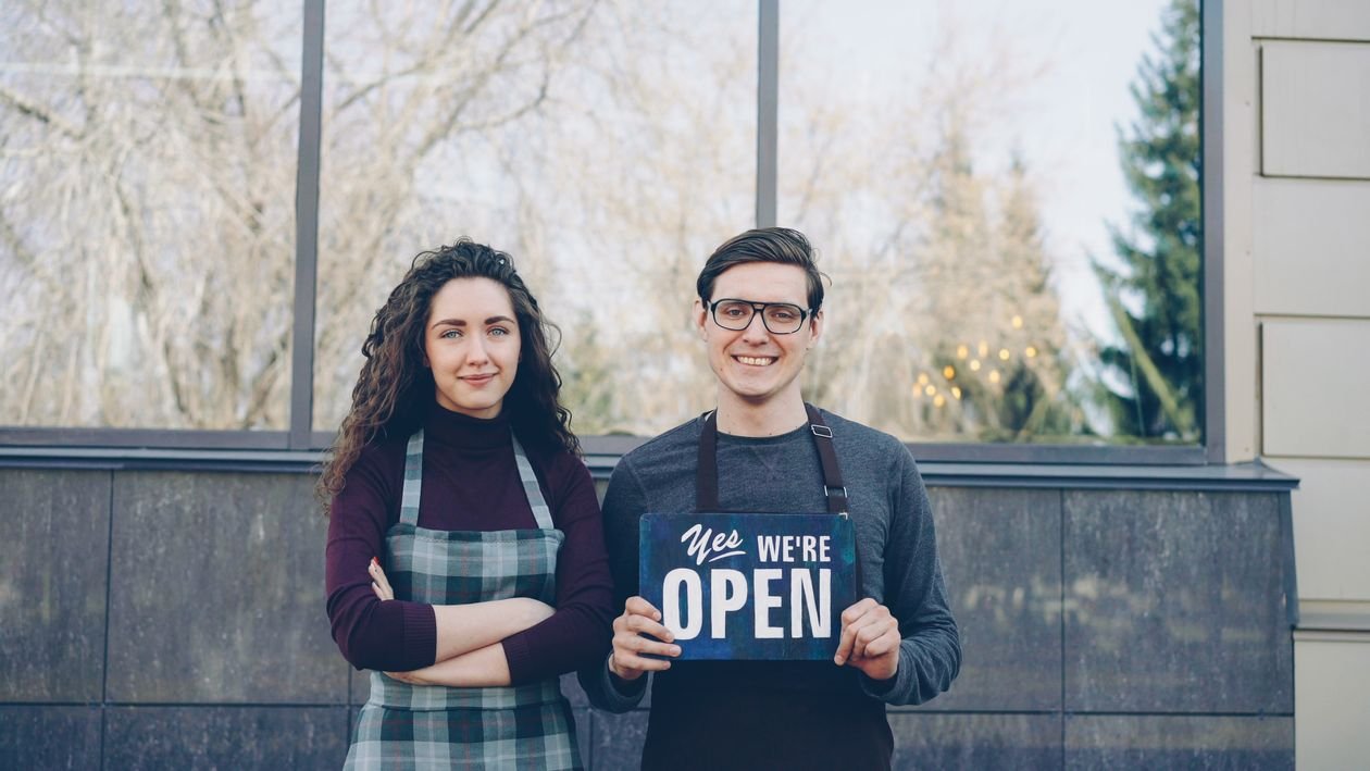 Two small business owners holding an open sign outside their shop